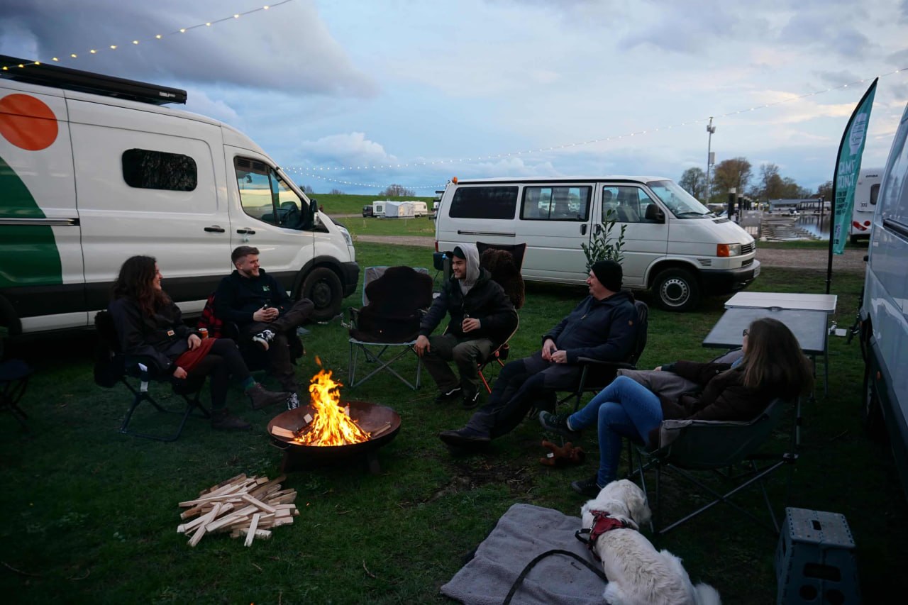 Reisende sitzen abends im Kreis um ein Feuer zwischen ihren Campern